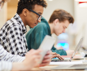 Estudante negro de cabelos curtos usando camisa xadrez escreve em seu laptop em sala com mais estudantes.
